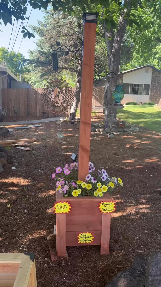 Tall redwood post with a solar lantern on top, a shepherd hook for a bird feeder, and a flower-filled planter box at the base.