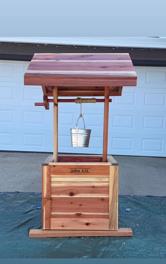 Finished cedar wishing well with a wide slab roof, rope-wrapped crank, galvanized bucket and John 4:14 nameplate.
