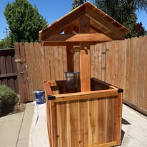 Tall cedar wishing well standing against a wood fence on a sunny patio.