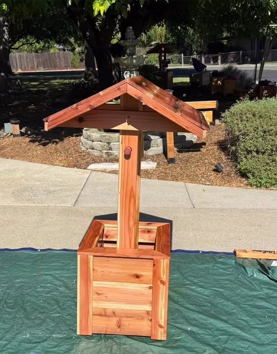 A freshly assembled wishing well with a steep peaked roof sitting on a tarp in the driveway, waiting to be stained.