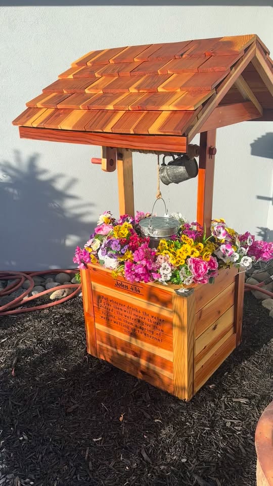Redwood wishing well planter with a hand-shingled roof, galvanized bucket on a rope crank, and the planter box overflowing with pink, purple and yellow blooms.